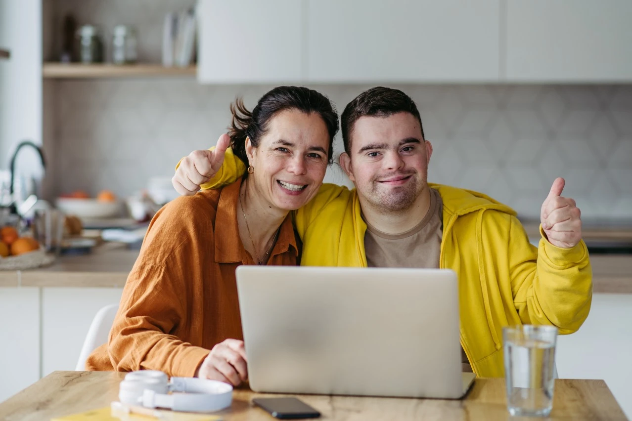 Happy disabled man with down syndrome sitting at table in front of laptop with support worker, both smiling at camera